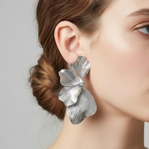以下が含まれることがあります： 銀色の花の形のステートメントイヤリング。イヤリングは、テクスチャ加工された、重ねられた花びらのデザインです。イヤリングは大きく、人目を引き、明るいメタリック仕上げです。イヤリングは人の耳に着用されています。
