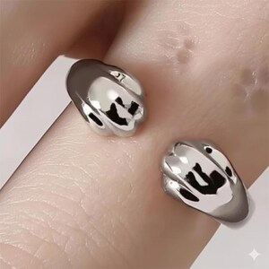 May include: A silver-colored ring with a unique design. The ring features two rounded, claw-like shapes with black accents. The ring is open, allowing for an adjustable fit. The ring is worn on a finger.