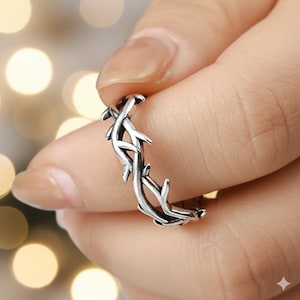 May include: A silver-colored ring with a detailed vine and leaf design. The ring is held in a hand, showcasing the intricate pattern of the jewelry. The ring's design features a repeating pattern of intertwined vines and leaves.