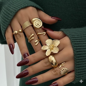 Pode incluir: Uma coleção de anéis dourados com vários designs, incluindo uma espiral, uma flor e uma concha. Os anéis são exibidos em uma mão com unhas longas de cor bordô, em um fundo verde.