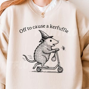 Könnte beinhalten: Beigefarbenes Sweatshirt mit dem Text "Off to cause a kerfuffle". Die Grafik zeigt eine Ratte mit Hexenhut, die Roller fährt und ein kleines Insekt jagt. Das Design ist in Schwarz und Weiß gehalten.