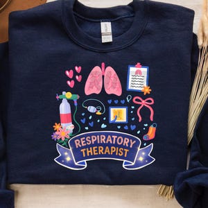 May include: Navy blue sweatshirt with a colorful graphic design. The design includes pink lungs, medical equipment, hearts, and the words "Respiratory Therapist" on a banner. A brown hat and dried flowers are in the background.