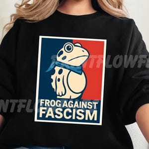 May include: Black sweatshirt featuring a graphic of a frog wearing a blue bandana, set against a red and blue background. The text "FROG AGAINST FASCISM" is displayed below the frog illustration in a white box.