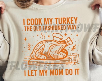 May include: Beige sweatshirt with orange graphic text and illustration. The text reads "I COOK MY TURKEY THE OLD FASHIONED WAY I LET MY MOM DO IT." The illustration depicts a cooked turkey with a chef's hat. A humorous Thanksgiving or holiday-themed design.