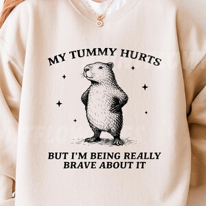 Op de afbeelding: Beige sweatshirt met een afbeelding van een capibara en de tekst "MY TUMMY HURTS BUT I'M BEING REALLY BRAVE ABOUT IT". De capibara staat met gekruiste poten. Vier zwarte sterren omringen de capibara.