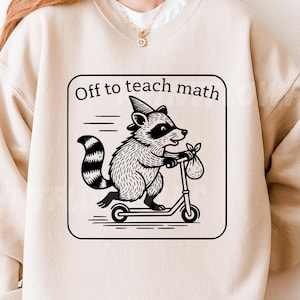 May include: Beige sweatshirt with a black and white graphic of a raccoon riding a scooter, wearing a party hat, and carrying a bag. The text "Off to teach math" is in a black rectangle.