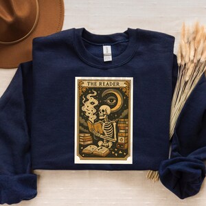 Puede incluir: Sudadera azul marino con un diseño de carta del tarot. La carta dice "THE READER" y muestra un esqueleto leyendo un libro, con humo, una luna creciente y un ojo. El diseño está enmarcado en un borde decorativo.