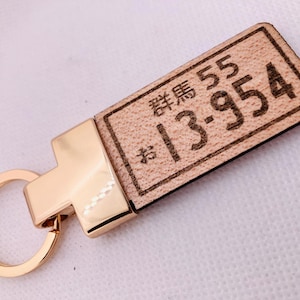 May include: A wooden keychain with a rectangular plate featuring engraved Japanese characters and numbers. The plate is attached to a gold-colored metal clasp and keyring. The text on the plate reads "群馬 55 13-954".