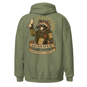 以下が含まれることがあります： オリーブグリーンのパーカー。軍服を着たアライグマのキャラクターが靴下を持っています。デザインには「SOCKRATESE」の文字と「CHANGE YOUR SOCKS. DRINK WATER. CARRY ON.」というフレーズが含まれています。