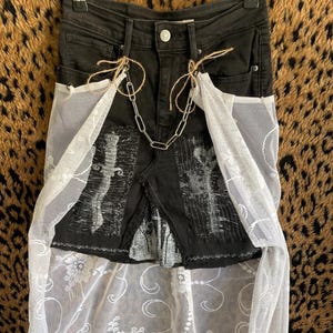 Könnte beinhalten: Schwarze Jeans mit weißem Spitzen-Overlay. Die Jeans haben eine Kette und dekorative Patches. Das Spitzen-Overlay hat ein Blumenmuster. Die Jeans hängen an einem Metallbügel.