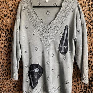 Könnte beinhalten: Ein hellgrauer Pullover mit V-Ausschnitt und langen Ärmeln. Der Pullover hat ein dekoratives Lochmuster um den Halsausschnitt und den Saum, mit rautenförmigen Aussparungen. Zwei schwarze Aufnäher mit weißen Illustrationen befinden sich auf der Vorderseite.