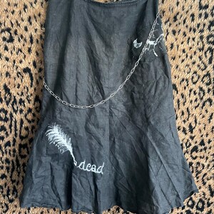 Könnte beinhalten: Schwarzer Jeansrock mit silberner Kette. Der Rock hat weiße, bemalte Designs und das Wort "dead". Der Rock hängt an einem Kleiderbügel.