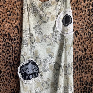 Könnte beinhalten: Ein langer, fließender Rock mit einem hellgrünen und beigefarbenen Paisley-Muster. Der Rock hat zwei dekorative Aufnäher: einen mit einem schwarz-weißen Motten-Design und einen mit einem schwarzen Kreis-Design. Der Rock hängt an einem Kleiderbügel.