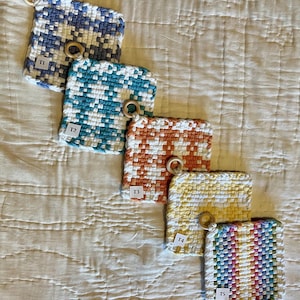 May include: A collection of five woven potholders in various colors, including blue, teal, orange, yellow, and a rainbow pattern. Each potholder has a wooden ring for hanging and a small white tag with a letter and number.