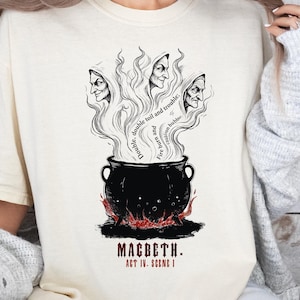 May include: Cream-colored t-shirt featuring a black and white illustration from Shakespeare's Macbeth. The design includes a cauldron with three witch faces emerging from smoke, with text that reads "Double, double toil and trouble". Below the cauldron, the text reads "Macbeth. Act IV. Scene I."