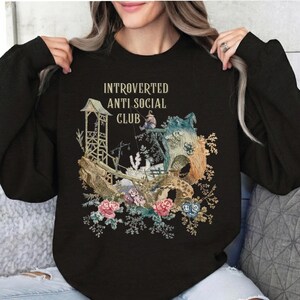Op de afbeelding: Een zwart sweatshirt met lange mouwen en een grafische print met de tekst "INTROVERTED ANTI SOCIAL CLUB" boven een illustratie van een klein figuurtje dat vist bij een houten constructie, omringd door gedetailleerde bloemmotieven.