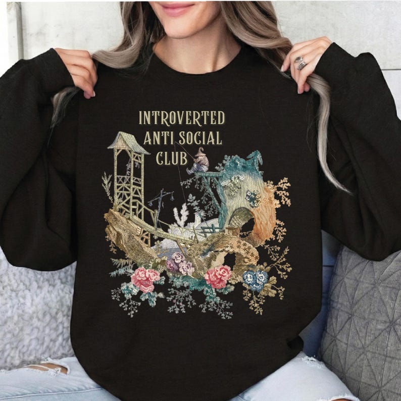 Op de afbeelding: Een zwart sweatshirt met lange mouwen en een grafische print met de tekst "INTROVERTED ANTI SOCIAL CLUB" boven een illustratie van een klein figuurtje dat vist bij een houten constructie, omringd door gedetailleerde bloemmotieven.