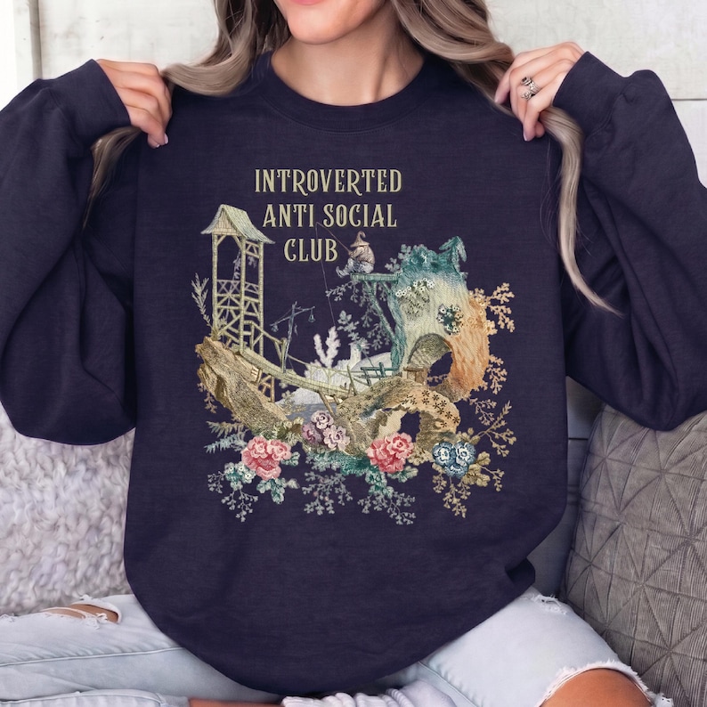 Op de afbeelding: Een donker marineblauw sweatshirt met ronde hals en een grafische print met de tekst "INTROVERTED ANTI SOCIAL CLUB" boven een illustratie van een houten structuur, een klein vissend figuurtje en kleurrijke bloemaccenten in roze en blauw.