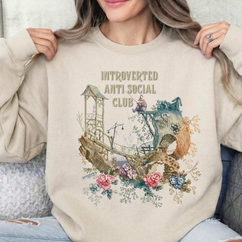 Op de afbeelding: Een beige sweater met ronde hals en een grafische print met de tekst "INTROVERTED ANTI SOCIAL CLUB" boven een illustratie van een figuur die vist vanaf een houten constructie op een klif, omringd door gedetailleerde bloemelementen.