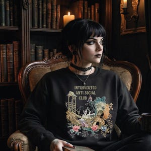 Op de afbeelding: Een zwart sweatshirt met lange mouwen en een grafische print met de tekst "INTROVERTED ANTI SOCIAL CLUB" boven een illustratie van een houten structuur, een klein figuurtje en kleurrijke bloemaccenten in roze, blauw en groen.