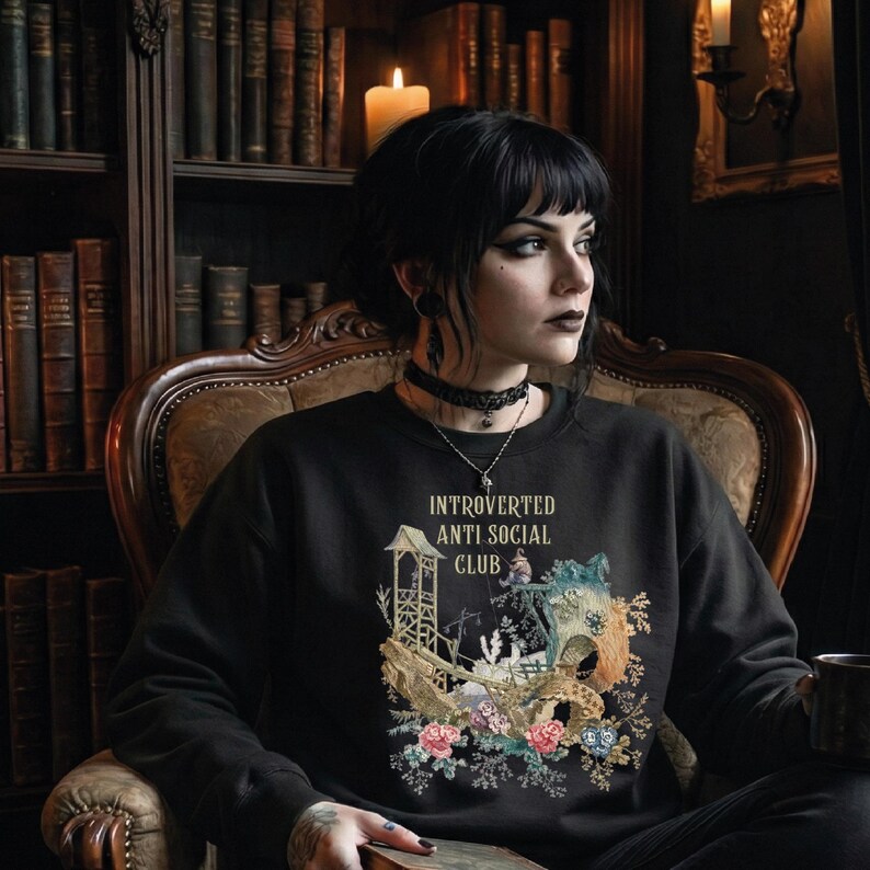 Op de afbeelding: Een zwart sweatshirt met lange mouwen en een grafische print met de tekst "INTROVERTED ANTI SOCIAL CLUB" boven een illustratie van een houten structuur, een klein figuurtje en kleurrijke bloemaccenten in roze, blauw en groen.