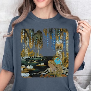 May include: A dark slate blue t-shirt featuring a graphic print of a toad sitting near a pond with water lilies. The scene includes tall, textured trees with hanging blue flowers and a bright blue butterfly resting on the toad's back.
