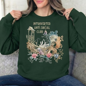 Op de afbeelding: Een donkergroene sweater met ronde hals en een grafische print met de tekst "INTROVERTED ANTI SOCIAL CLUB" boven een illustratie van een houten structuur, een klein figuurtje en bloemelementen in roze, blauwe en bruine tinten.