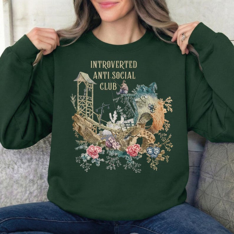 Op de afbeelding: Een donkergroene sweater met ronde hals en een grafische print met de tekst "INTROVERTED ANTI SOCIAL CLUB" boven een illustratie van een houten structuur, een klein figuurtje en bloemelementen in roze, blauwe en bruine tinten.