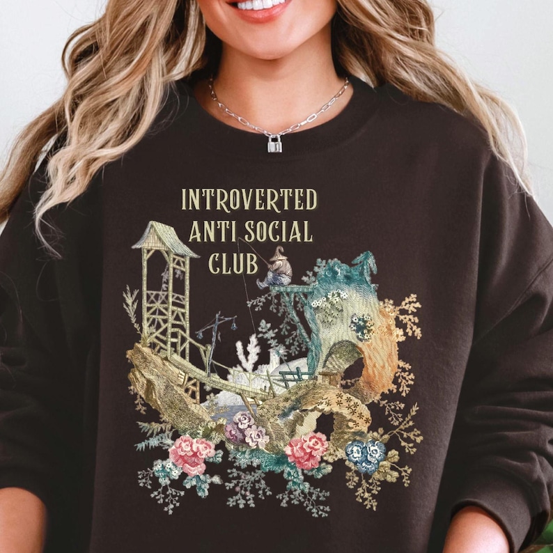 Op de afbeelding: Een donkerbruin sweatshirt met ronde hals en een grafische print met de tekst "INTROVERTED ANTI SOCIAL CLUB" boven een illustratie van een klein figuurtje dat vist bij een houten constructie, een rotsachtig landschap en diverse bloemen.