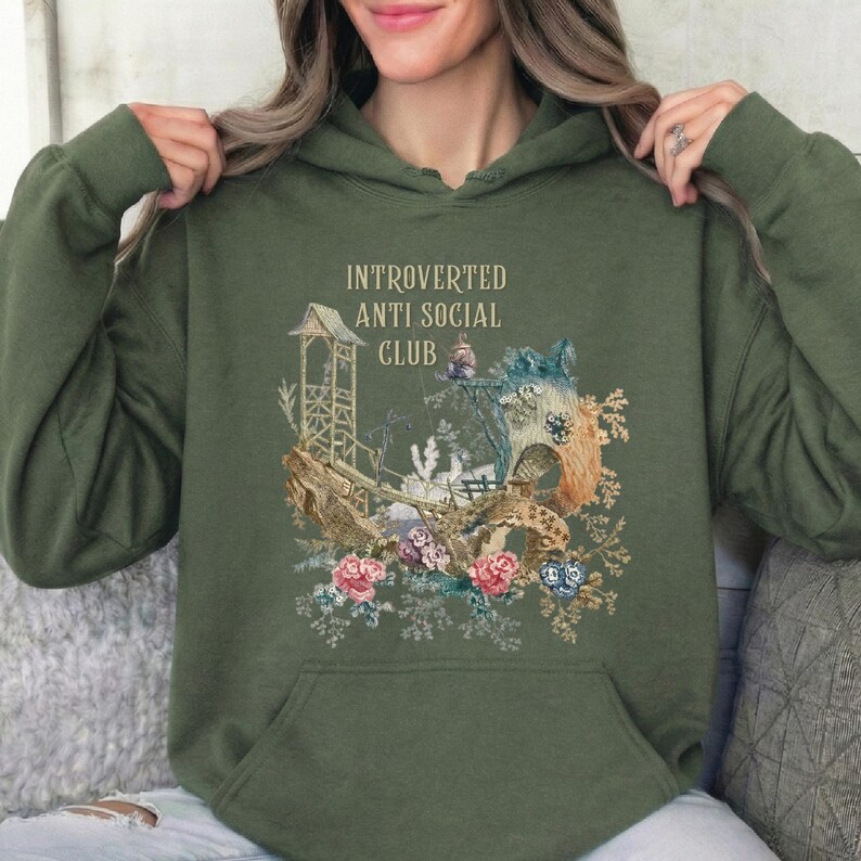 Op de afbeelding: Een olijfgroene hoodie met een grafische print met de tekst "INTROVERTED ANTI SOCIAL CLUB" boven een illustratie van een houten structuur, een brug en diverse kleurrijke bloemen in tinten roze, blauw en bruin.