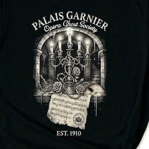 May include: Black long-sleeve shirt featuring a detailed graphic of a candelabra with lit candles, roses, and sheet music. The text "Palais Garnier Opera Ghost Society" and "EST. 1910" are also displayed. The design has a vintage, gothic aesthetic.