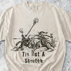 May include: A cream-colored crew neck t-shirt featuring a black ink-style illustration of two armored figures jousting on horseback. Below the graphic, the text reads "Tis But A Scratch" with a horizontal line drawn through the word Scratch.