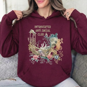 Op de afbeelding: Een bordeauxrode hoodie met de tekst INTROVERTED ANTI SOCIAL CLUB in gouden letters. De afbeelding toont een houten constructie, een grote boom en kleurrijke florale elementen, waaronder roze en blauwe rozen aan de onderkant.