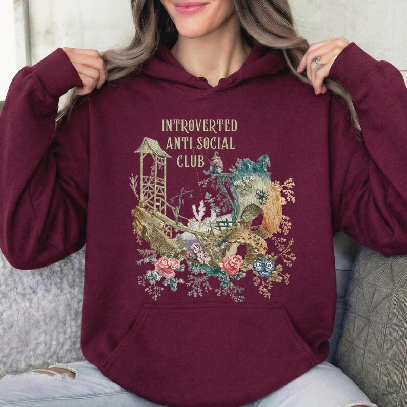 Op de afbeelding: Een bordeauxrode hoodie met de tekst INTROVERTED ANTI SOCIAL CLUB in gouden letters. De afbeelding toont een houten constructie, een grote boom en kleurrijke florale elementen, waaronder roze en blauwe rozen aan de onderkant.