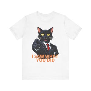 以下が含まれることがあります： 黒いスーツと赤いネクタイを身に着けた黒猫のグラフィックがプリントされた白いTシャツ。猫は驚いた表情で前を指差している。猫の下には、オレンジ色の文字で「I SAW WHAT YOU DID」と書かれている。