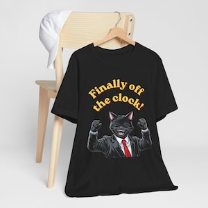 以下が含まれることがあります： 黒の半袖Tシャツ。スーツと赤いネクタイを着用し、両拳を突き上げた黒猫のグラフィックが描かれている。猫の上部には、暗い縁取りが施された黄色の文字で「Finally off the clock!」と記されている。