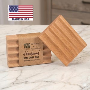 May include: Two handcrafted hardwood soap saver dishes. One dish is upright, the other is lying down. The dishes are made of light brown wood with a grooved design. The label reads "Wild Herb Hardwood Soap Saver Dish". The American flag is in the background.