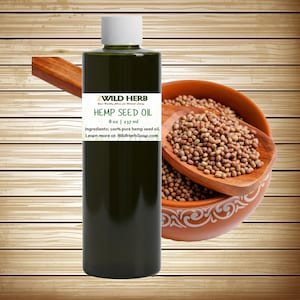 May include: A dark green bottle of Wild Herb Hemp Seed Oil, 8 oz (237 ml), with a white cap. The label includes the text "Ingredients: 100% pure hemp seed oil." A wooden bowl filled with hemp seeds and a wooden spoon are in the background.