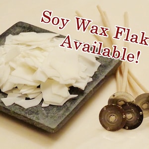 Pode incluir: Flocos de cera de soja brancos sobre uma superf&iacute;cie de pedra cinza, ao lado de um pacote de pavios de vela brancos e quatro adesivos de pavios de vela prateados. O texto "Soy Wax Flakes Available!" est&aacute; escrito em letras vermelhas sobre fundo branco.