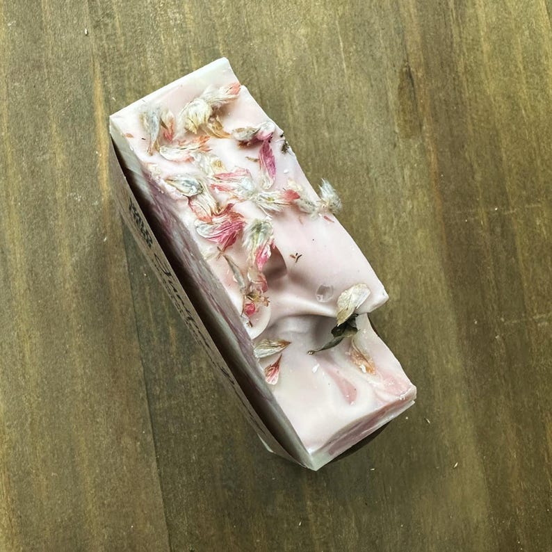 May include: A rectangular bar of pink soap with dried floral accents. The soap has a marbled appearance and is partially wrapped in a paper sleeve. The dried flowers are in shades of pink, white, and green.
