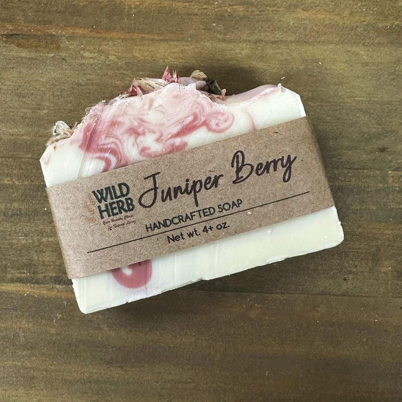 May include: A handcrafted Juniper Berry soap bar with a white base and pink marbling. The soap is wrapped with a brown paper band that reads "Wild Herb Handcrafted Soap" and "Net wt. 4+ oz."