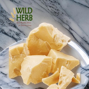May include: Close-up of broken pieces of pale yellow shea butter in a white bowl. The bowl is on a marble surface. The text "WILD HERB Your Healthy Choice for Natural Living" is in the upper left corner of the image.