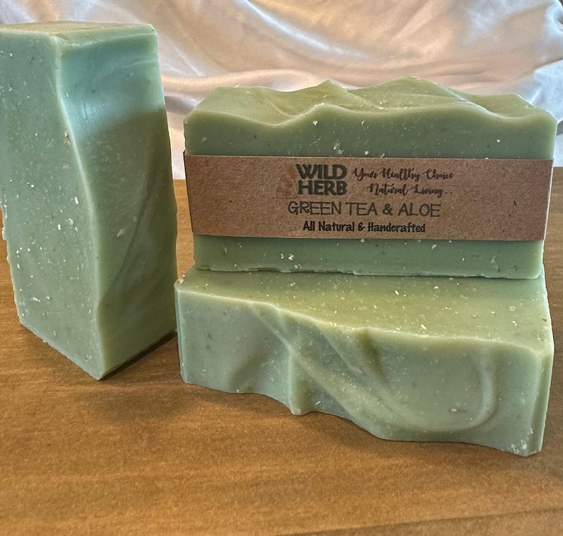 May include: Three bars of green tea and aloe soap, made with natural ingredients and handcrafted. The soap is wrapped in brown paper with the text "WILD HERB" and "Green Tea & Aloe" printed on it.