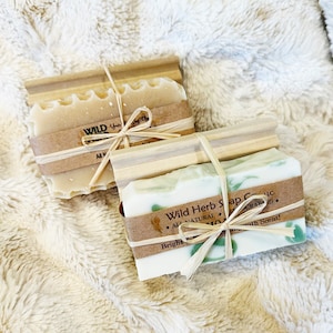 May include: Two bars of handmade soap on wooden soap dishes. The soap on the left is a plain white bar with a brown label that says "Wild Herb" and "All Natural Handcrafted". The soap on the right is a white bar with green swirls and a brown label that says "Wild Herb Soap Co. LLC", "All Natural Handcrafted", "Bright", and "with Scent!"