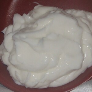 Pode incluir: Um close-up de uma subst&acirc;ncia branca e cremosa, provavelmente iogurte ou creme de leite, em uma tigela vermelha. A subst&acirc;ncia cremosa &eacute; lisa e tem uma superf&iacute;cie ligeiramente texturizada.