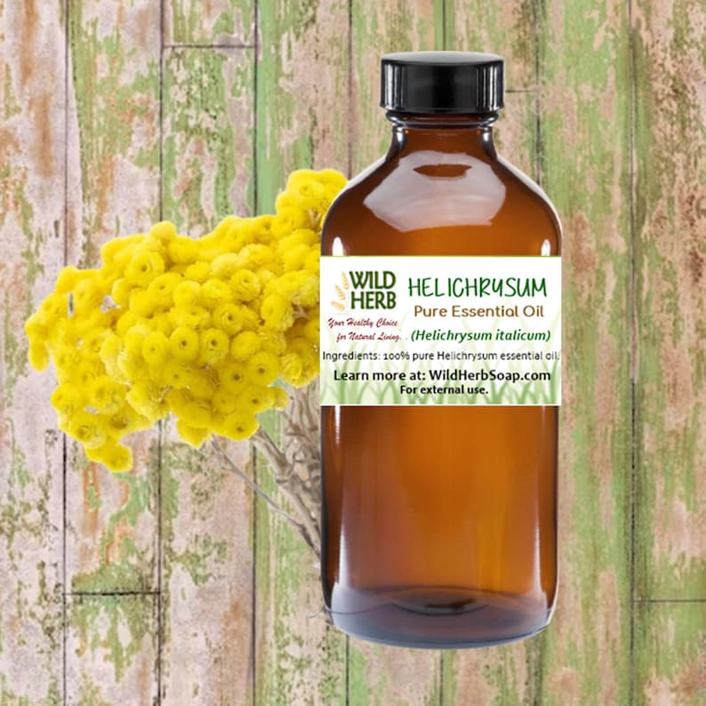 May include: A brown glass bottle with a label that reads "WILD HERB HELICHRYSUM Pure Essential Oil (Helichrysum italicum)". The label also includes the text "Your Healthy Choice for Natural Living" and "Ingredients: 100% pure Helichrysum essential oil". The bottle is sitting on a wooden surface next to a bunch of yellow flowers.
