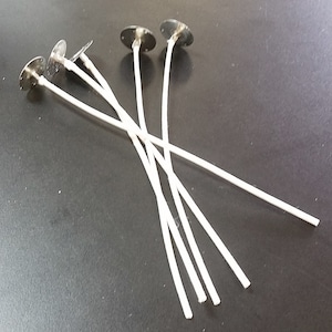 May include: A collection of candle wicks with metal bases. The wicks are white, and the metal bases are silver. The wicks are arranged in a scattered pattern on a dark surface. These are candle-making supplies.