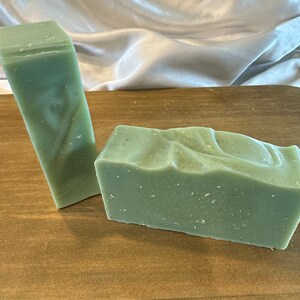 May include: Two bars of green soap on a wooden surface. One bar is rectangular and the other is a long, thin rectangle.