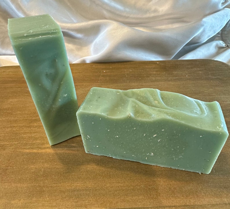May include: Two bars of green soap on a wooden surface. One bar is rectangular and the other is a long, thin rectangle.