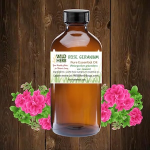 May include: A brown glass bottle with a black cap, labeled "WILD ROSE GERANIUM HERB Pure Essential Oil (Pelargonium graveolens var. roseum)". The label also states "Your Healthy Choice for Natural Living" and "Ingredients: 100% Rose Geranium essential oil." The bottle is sitting on a wooden surface with pink flowers in the background.